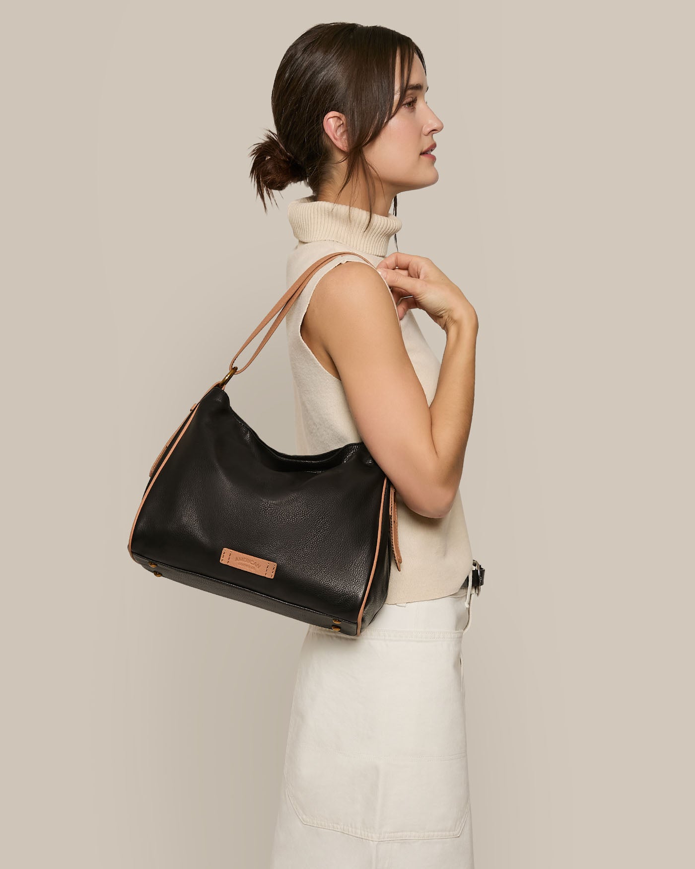 A person with tied-back hair in profile wears a sleeveless beige turtleneck and a white skirt. They hold the adjustable strap of an American Leather Co. Dean Double Entry Convertible Hobo with tan accents over their shoulder, set against a plain beige background.