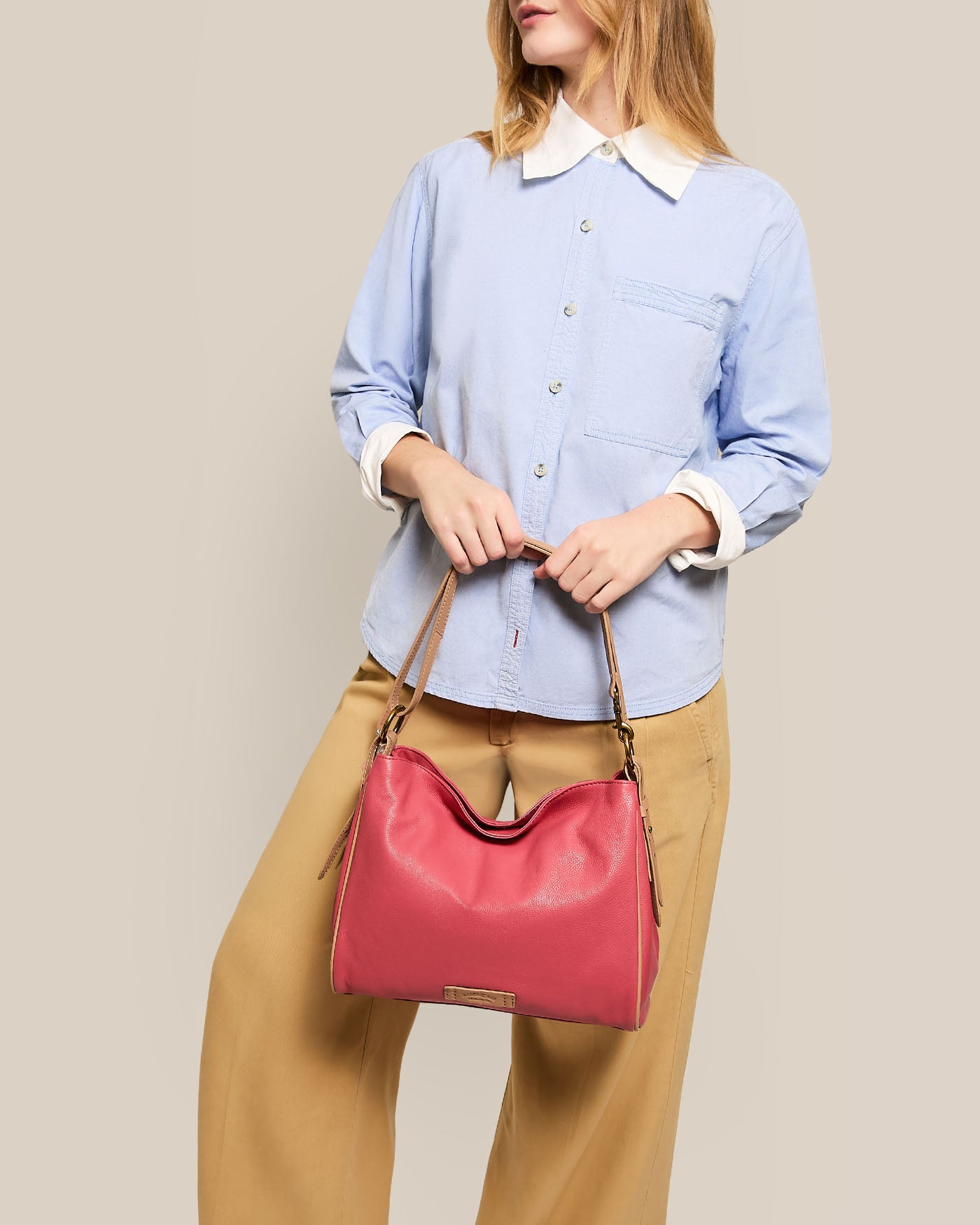 A person wearing a light blue button-up shirt and beige pants holds an American Leather Co. Dean Double Entry Convertible Hobo with an adjustable strap against a neutral beige background.