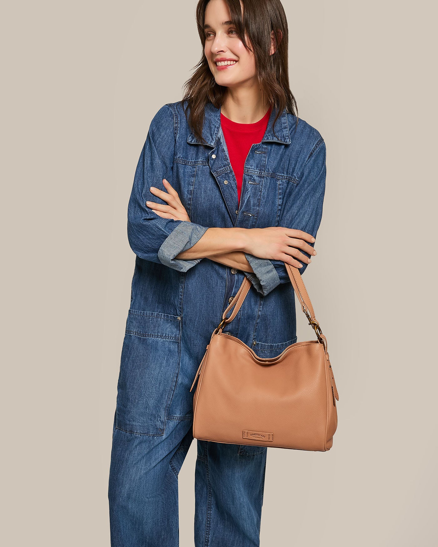 A person in a denim jumpsuit and red shirt beams while holding the stylish Dean Double Entry Convertible Hobo by American Leather Co. The tan handbag with an adjustable strap elevates the elegant simplicity of the plain beige backdrop.