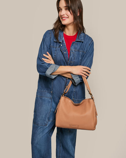 A person in a denim jumpsuit and red shirt beams while holding the stylish Dean Double Entry Convertible Hobo by American Leather Co. The tan handbag with an adjustable strap elevates the elegant simplicity of the plain beige backdrop.