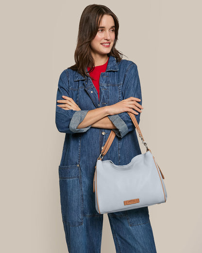 A woman with long brown hair in a denim outfit and red shirt poses with her American Leather Co. Dean Double Entry Convertible Hobo, featuring an adjustable strap, over her shoulder against a plain beige background.