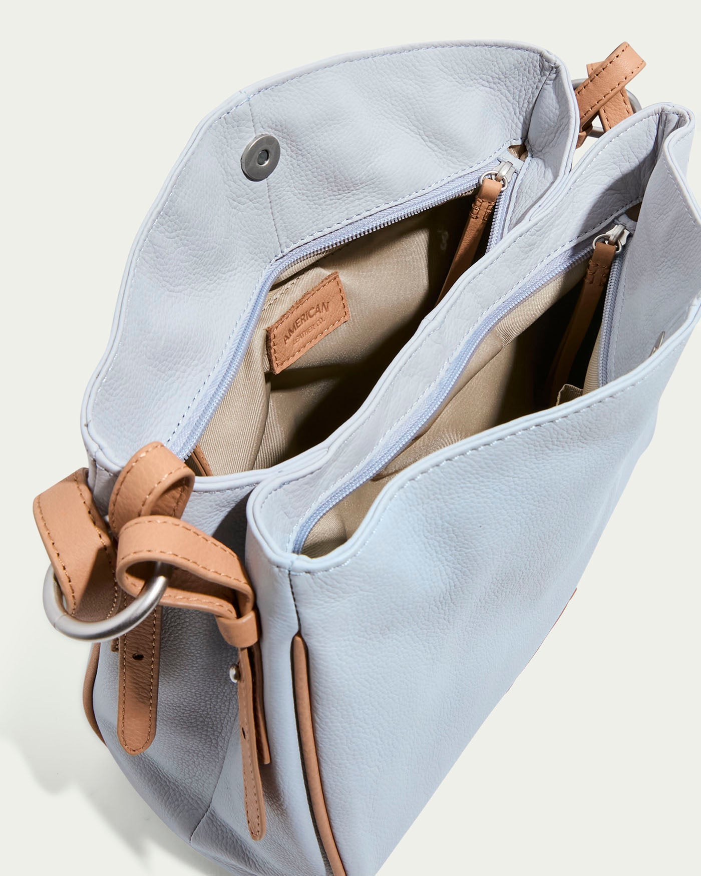 Unfasten the Dean Double Entry Convertible Hobo by American Leather Co., a light blue bag with tan glove leather straps. It has a beige interior with multiple pockets, snap closure, and an adjustable strap for easy organizing and carrying.