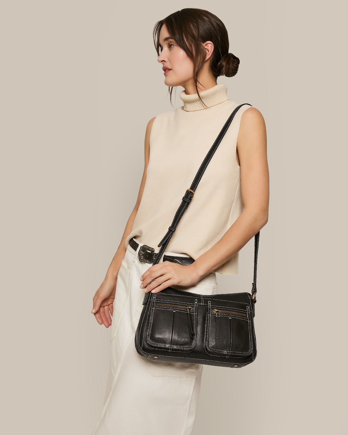 A person in a sleeveless beige turtleneck and white skirt carries the spacious American Leather Co. Holton Crossbody bag, featuring two front pockets crafted from glove leather, while posing against a plain light background.