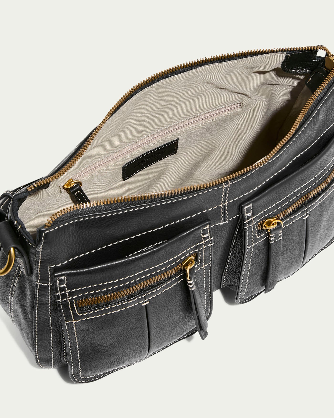 The American Leather Co. Holton Crossbody is a black glove leather bag with two front zippered pockets, elegant white stitching, an open main compartment with a beige interior and inner zippered pocket, and gold hardware including zippers and side rings, making it spacious for any occasion.