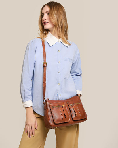 A person wearing a light blue button-up shirt and beige pants stands against a neutral background, carrying the Holton Crossbody by American Leather Co., crafted from glove leather with an adjustable strap. Their hair is styled in loose waves.