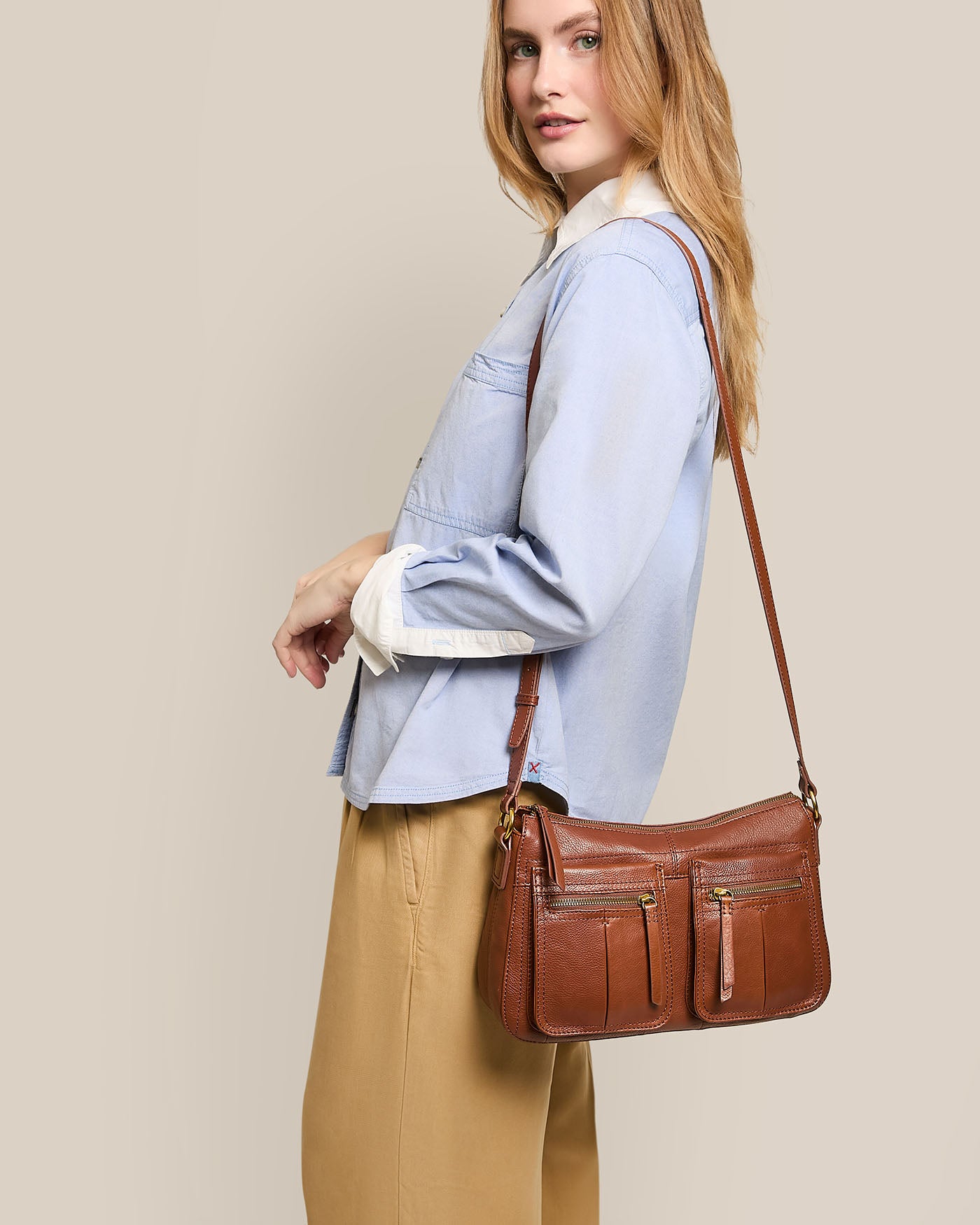 A person in a light blue shirt and tan pants holds an American Leather Co. Holton Crossbody, crafted from glove leather. The brown leather bag features multiple zippered pockets and an adjustable strap, standing out against the plain beige background.