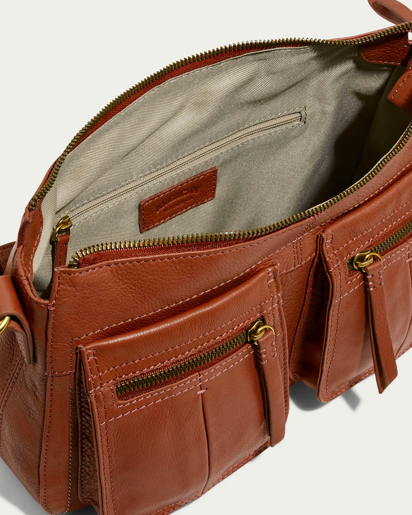 The Holton Crossbody by American Leather Co. is made from rich brown glove leather, featuring two front zippered pockets, an open main compartment with light beige lining, a zippered inner pocket, gold-tone hardware, and an adjustable strap for elegant functionality.