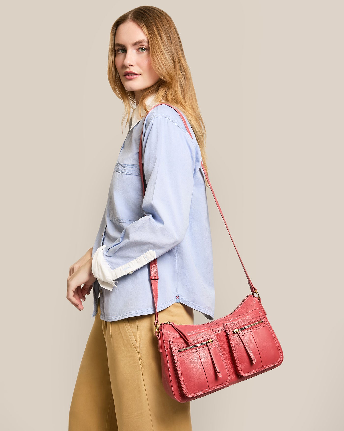 Against a plain background, a woman in a light blue shirt and tan pants carries the Holton Crossbody by American Leather Co., made of glove leather with multiple pockets and an adjustable strap.