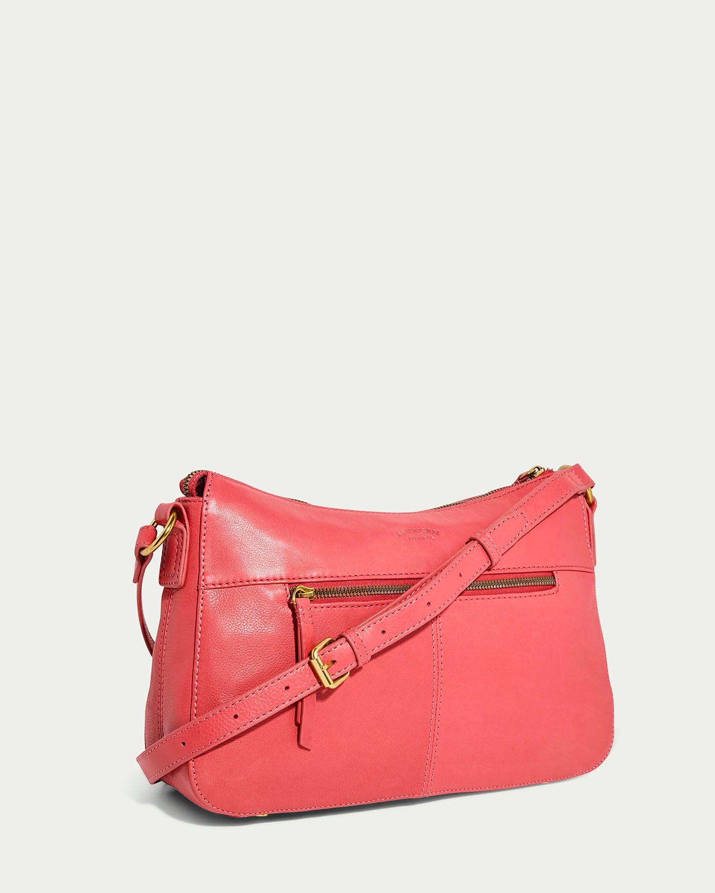 The Holton Crossbody by American Leather Co. is a stylish glove leather bag in coral, with an adjustable strap, gold buckles, and a sleek texture. Its front zipper pocket adds charm as it sits angled against a plain, light background.