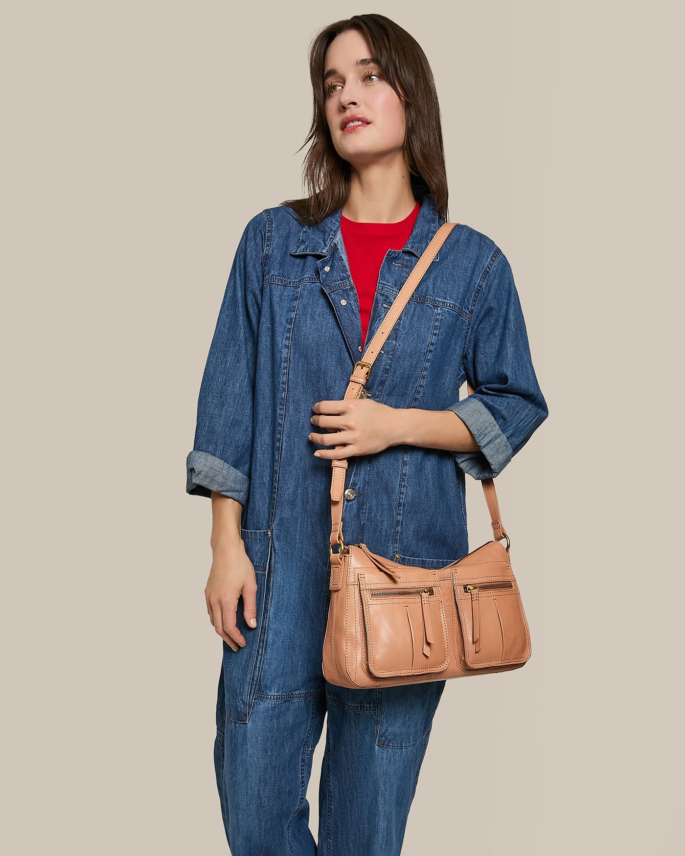 A woman in a denim jumpsuit and red shirt holds the Holton Crossbody by American Leather Co., a tan glove leather bag with an adjustable strap, standing against a plain beige background.