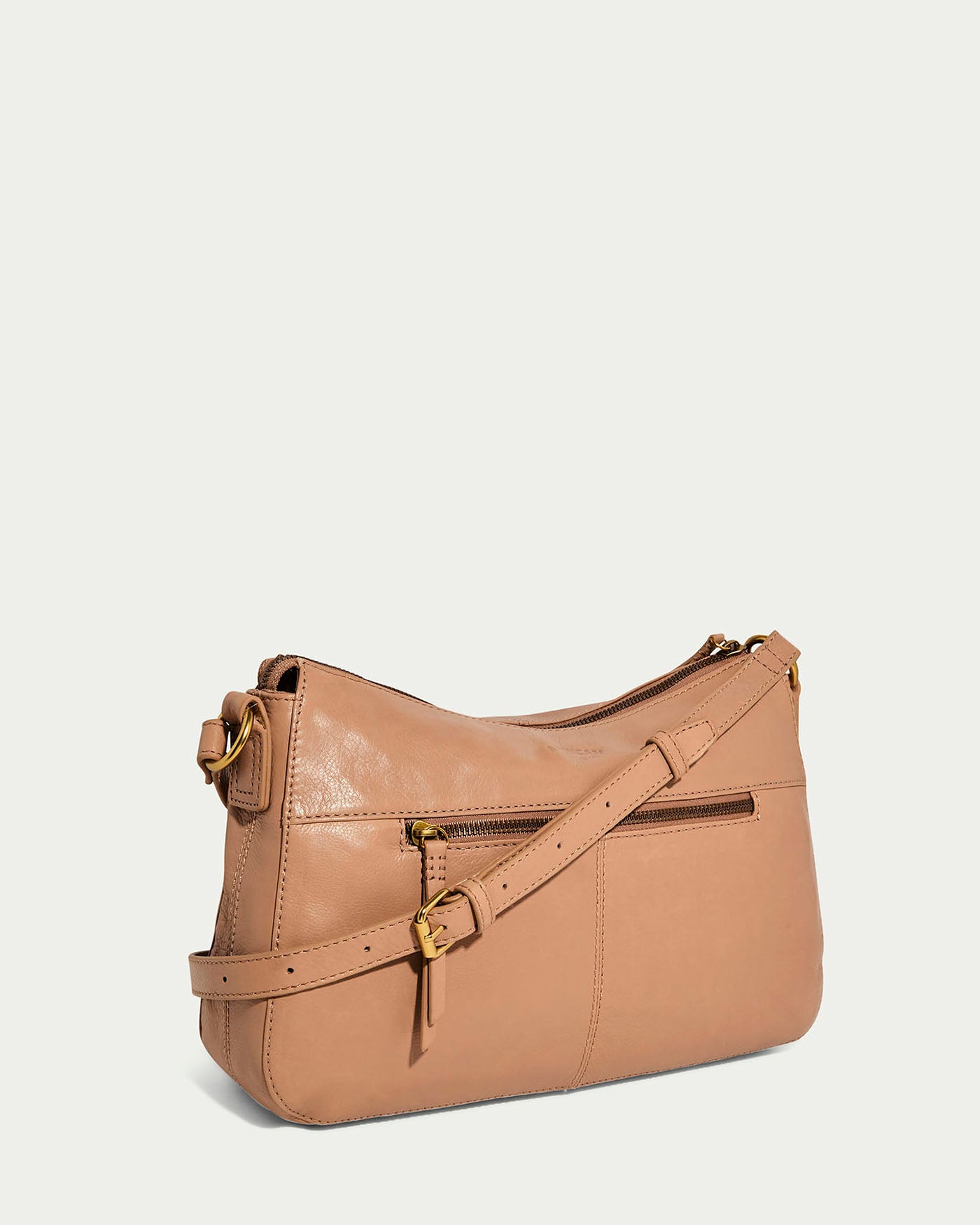 The American Leather Co. Holton Crossbody is a tan leather shoulder bag made from supple glove leather, featuring a curved top, front zipper pocket, and an adjustable strap with a gold buckle, elegantly set against a light gray background.