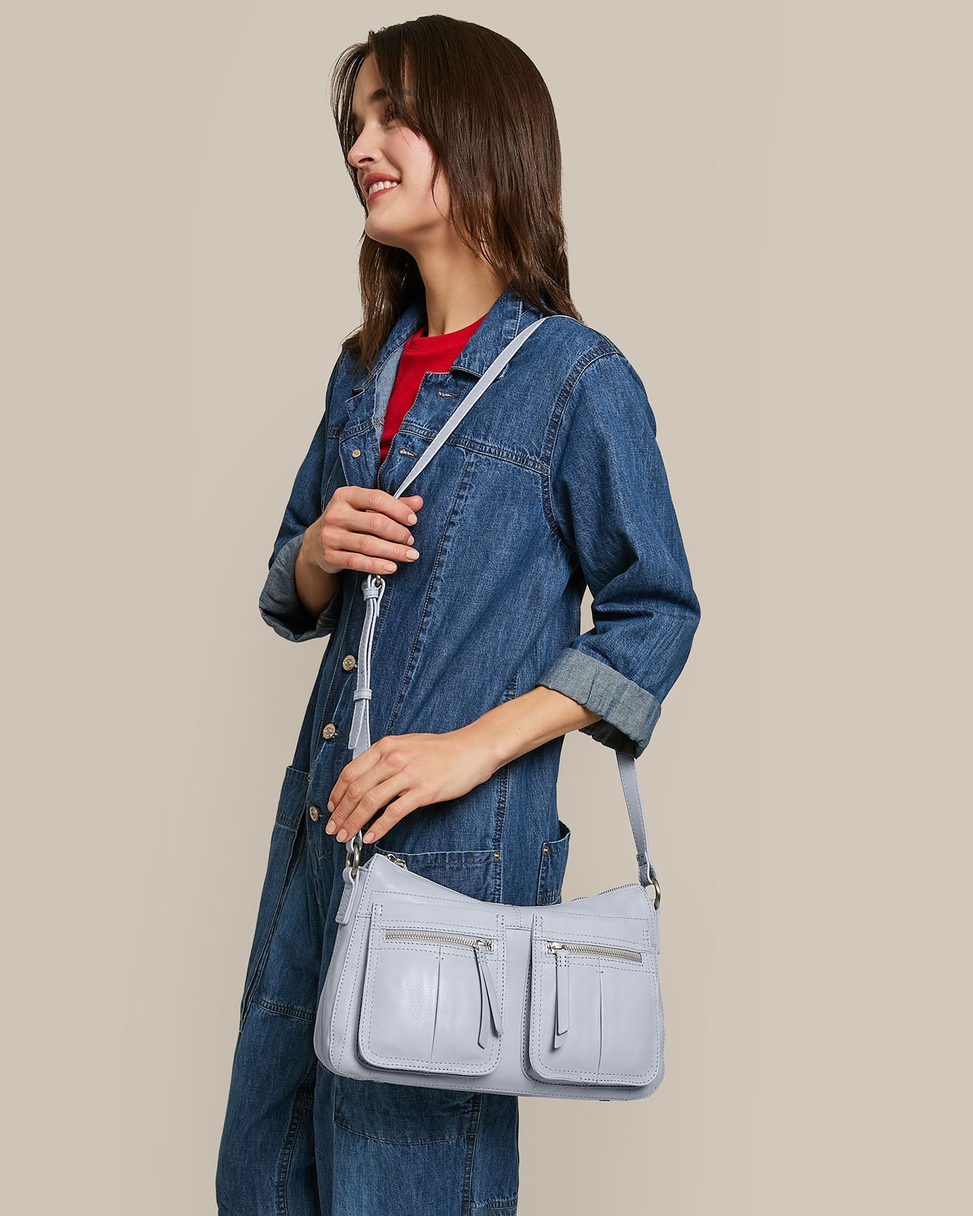 A woman smiles, holding the Holton Crossbody by American Leather Co., a light blue glove leather bag with multiple pockets and an adjustable strap. She's in a denim jumpsuit and red shirt against a solid beige background.