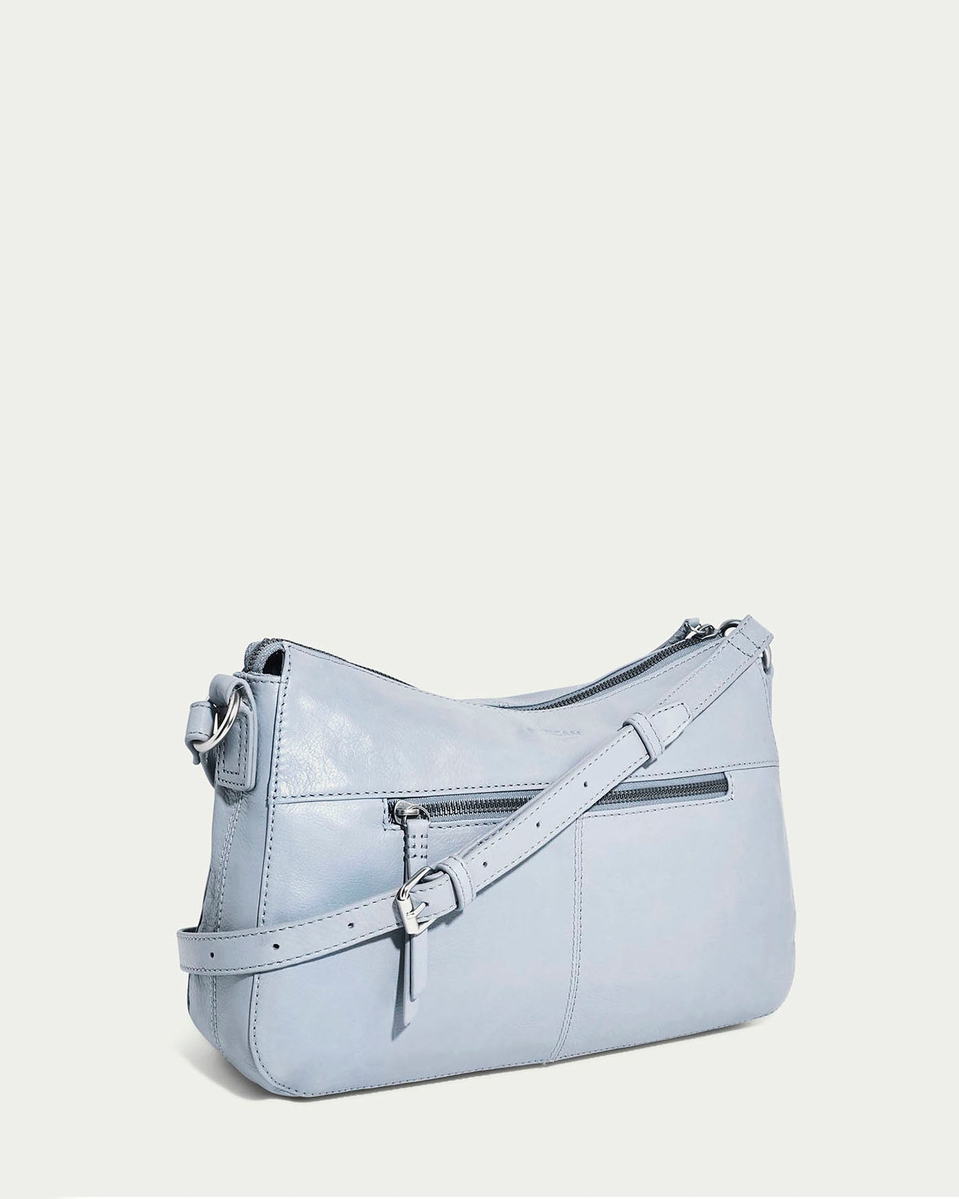 The American Leather Co.'s Holton Crossbody, a light blue glove leather handbag, is set against a plain background. It has an adjustable strap, front zip pocket, and minimal stitching for a sleek, modern look.