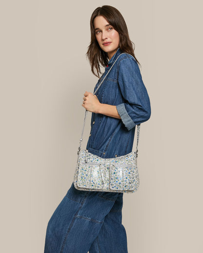 A person in a denim jumpsuit stands sideways, smiling against a beige background. With long brown hair, they hold the American Leather Co.’s Holton Crossbody—a white bag with multicolored speckles, glove leather details, and multiple pockets.