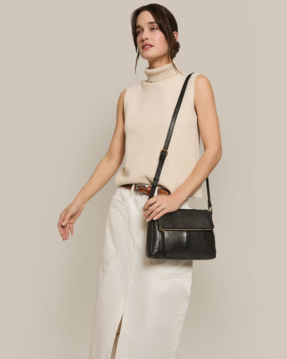 Wearing a sleeveless beige turtleneck and white skirt with a front slit, the person elegantly holds the Lee Double Entry Crossbody by American Leather Co., crafted from genuine black leather. Its adjustable strap offers both comfort and versatility against a neutral background.