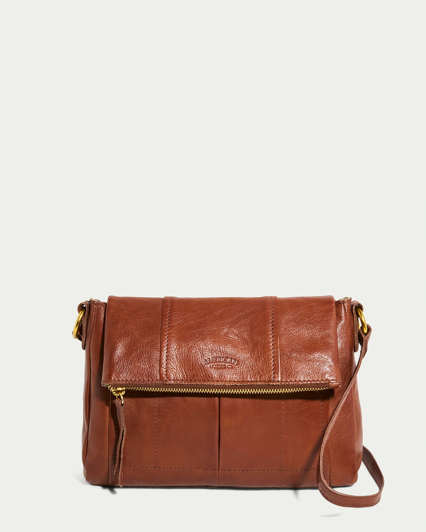 The Lee Double Entry Crossbody by American Leather Co. in Brandy features a flap closure, a front gold zipper pocket, and small gold clasps for the adjustable strap. It stands out against a plain, light-colored background.