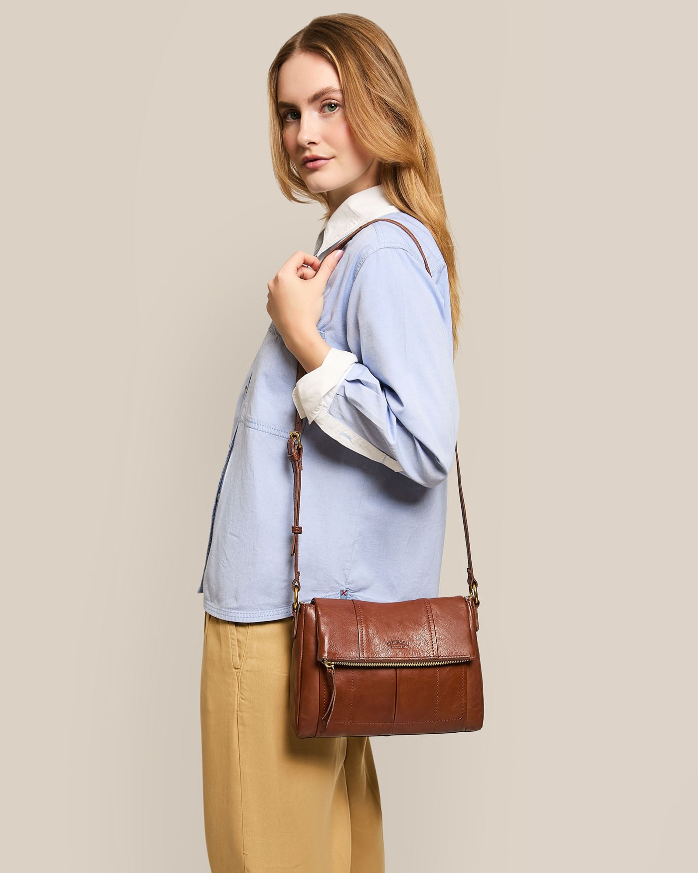 A person with long hair stands sideways, wearing a light blue shirt and beige pants, holding an American Leather Co. Lee Double Entry Crossbody bag featuring an adjustable strap made of genuine leather against a plain beige background.