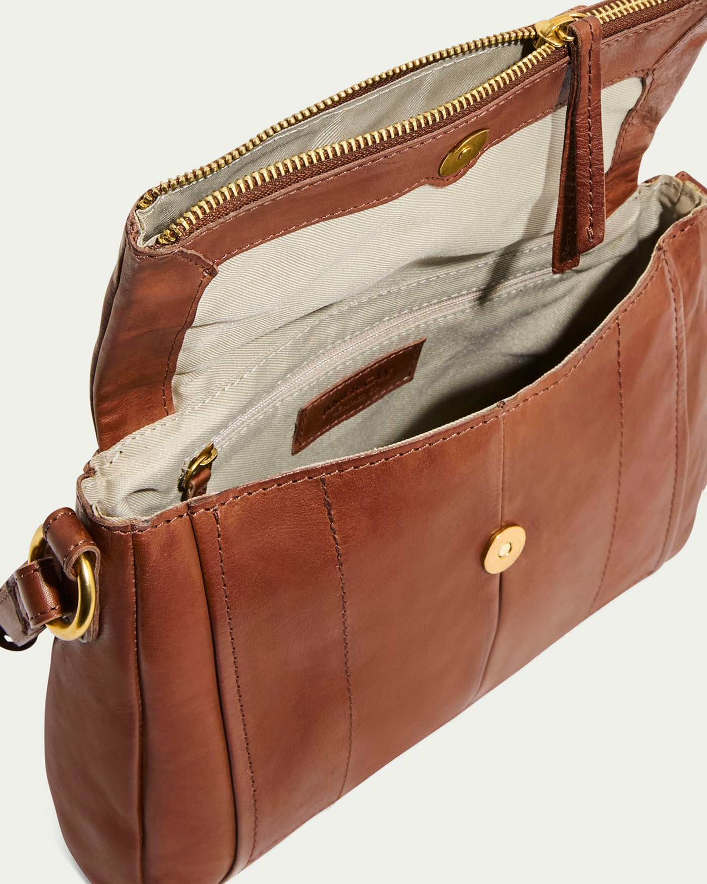 The Lee Double Entry Crossbody by American Leather Co. is an open brown genuine leather handbag with a gold zipper and button closure. It features a beige lining, multiple compartments including a zippered pocket, and an adjustable strap with a gold ring for versatile crossbody wear.