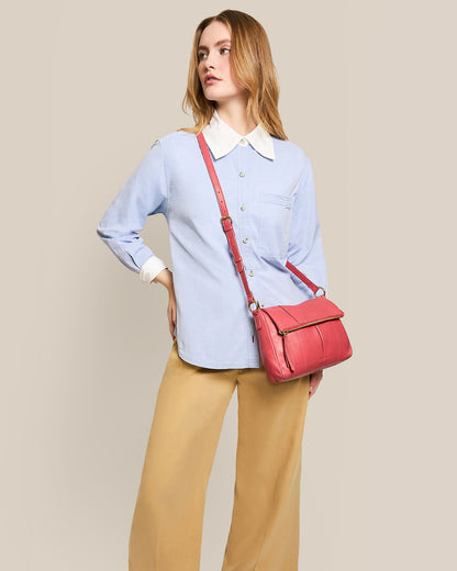 A woman stands against a plain background, wearing a light blue button-up shirt with white cuffs and collar, beige pants, and the American Leather Co. Lee Double Entry Crossbody in red genuine leather. Her head is slightly turned to the side.