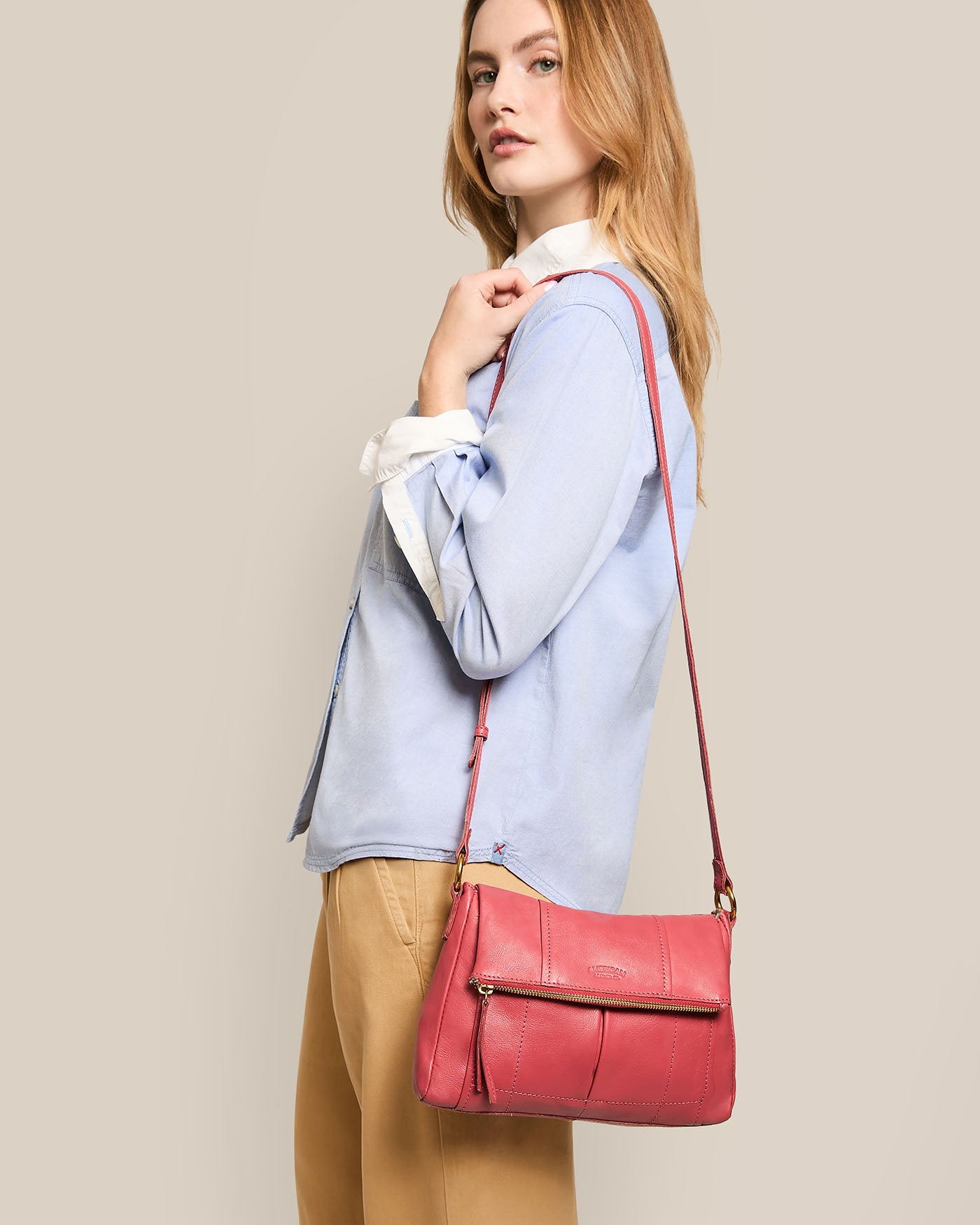 A woman with long hair, dressed in a light blue shirt and beige pants, poses with her left hand on her chin. She carries an American Leather Co. Lee Double Entry Crossbody bag against a plain beige background.