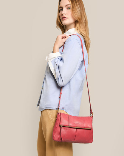 A woman with long hair, dressed in a light blue shirt and beige pants, poses with her left hand on her chin. She carries an American Leather Co. Lee Double Entry Crossbody bag against a plain beige background.