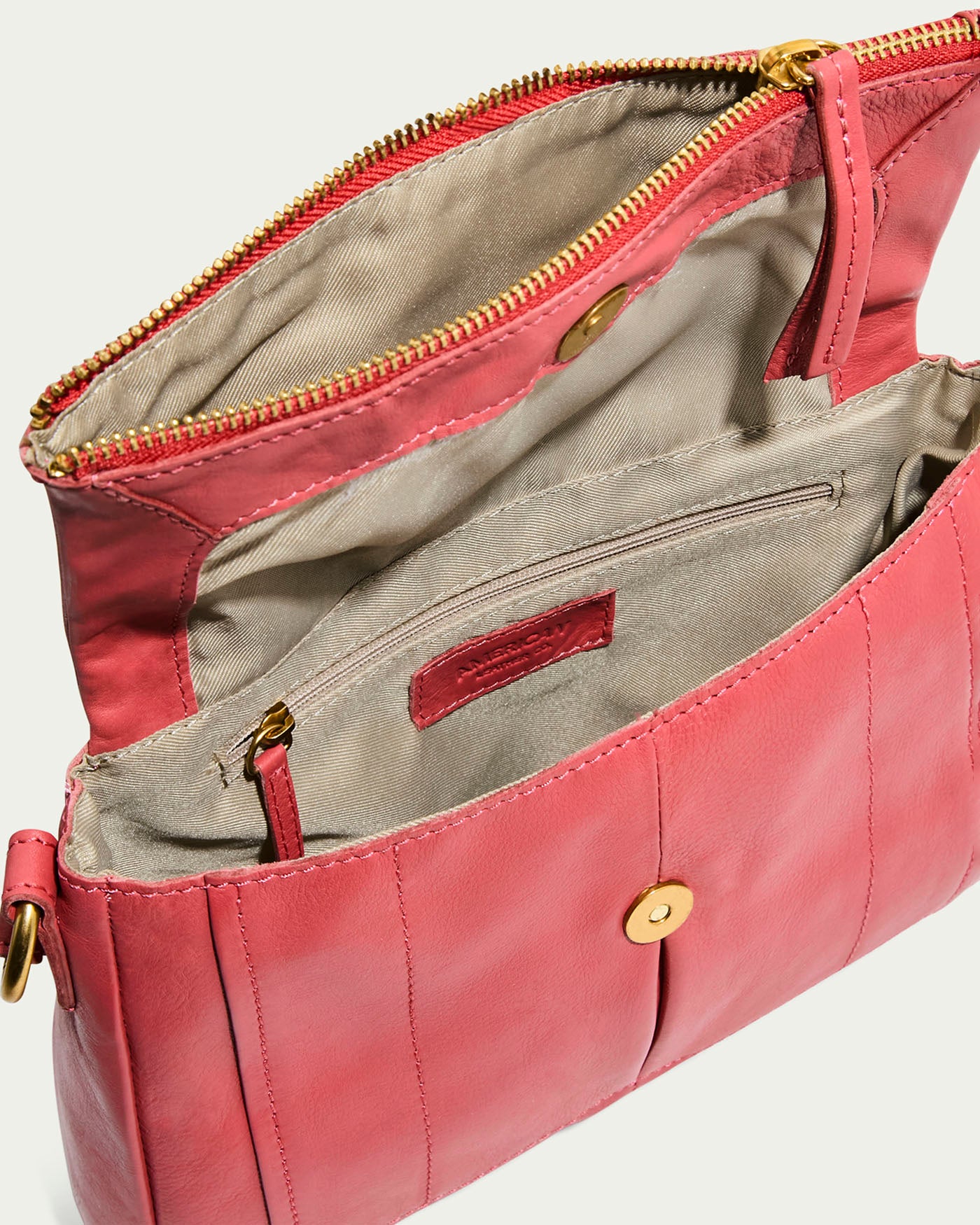 The Lee Double Entry Crossbody by American Leather Co. is a pink genuine leather handbag with gold hardware and a beige interior, featuring multiple zippered and buttoned compartments for organized storage. It includes an adjustable strap for transforming into a versatile crossbody bag.