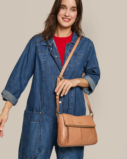 A person in a denim jumpsuit and red shirt smiles holding the Lee Double Entry Crossbody by American Leather Co. against a beige background.