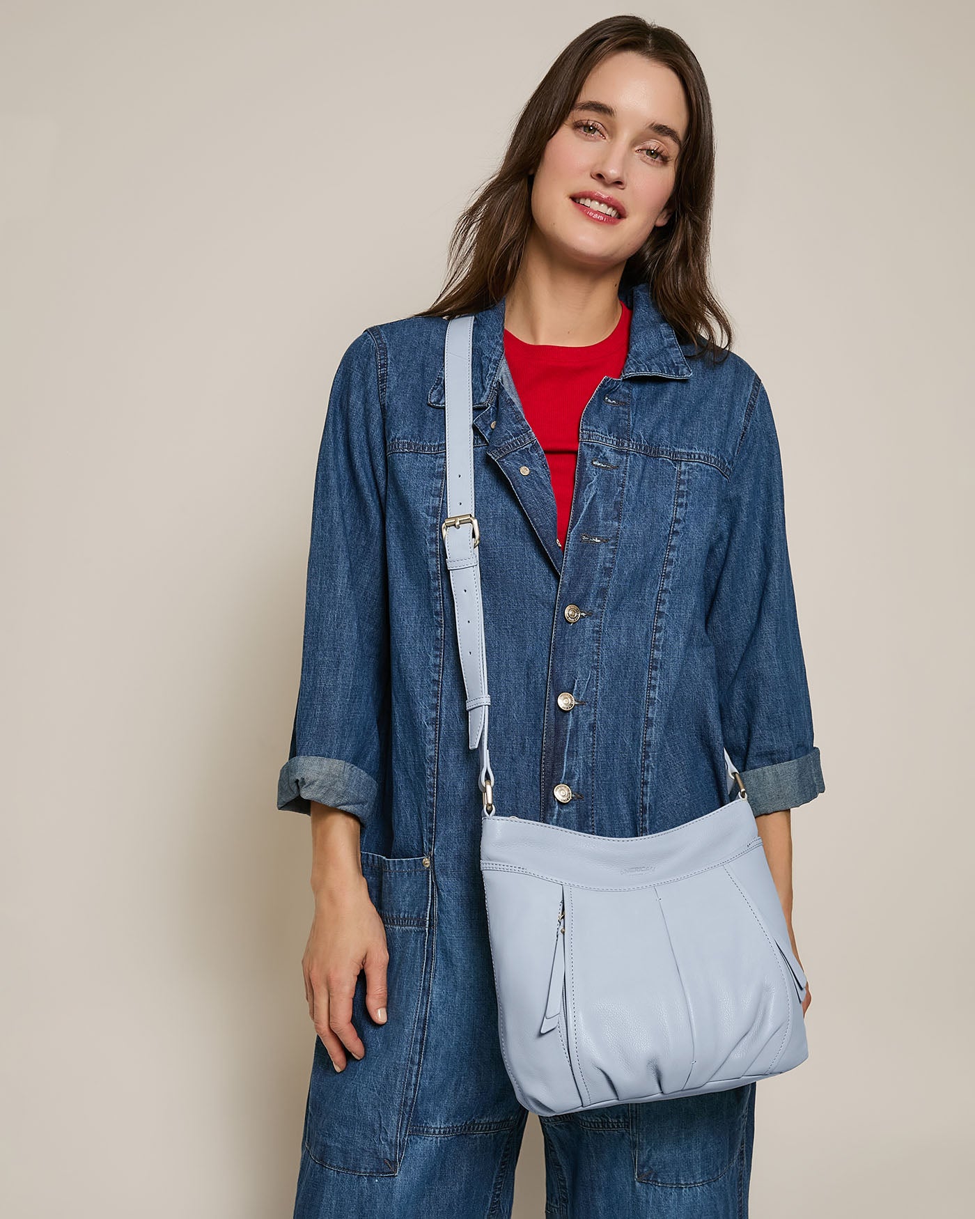 A person in a denim jumpsuit and red top stands against a plain background, smiling while holding an American Leather Co. Lennie Double Entry Crossbody bag in light blue with their right hand.