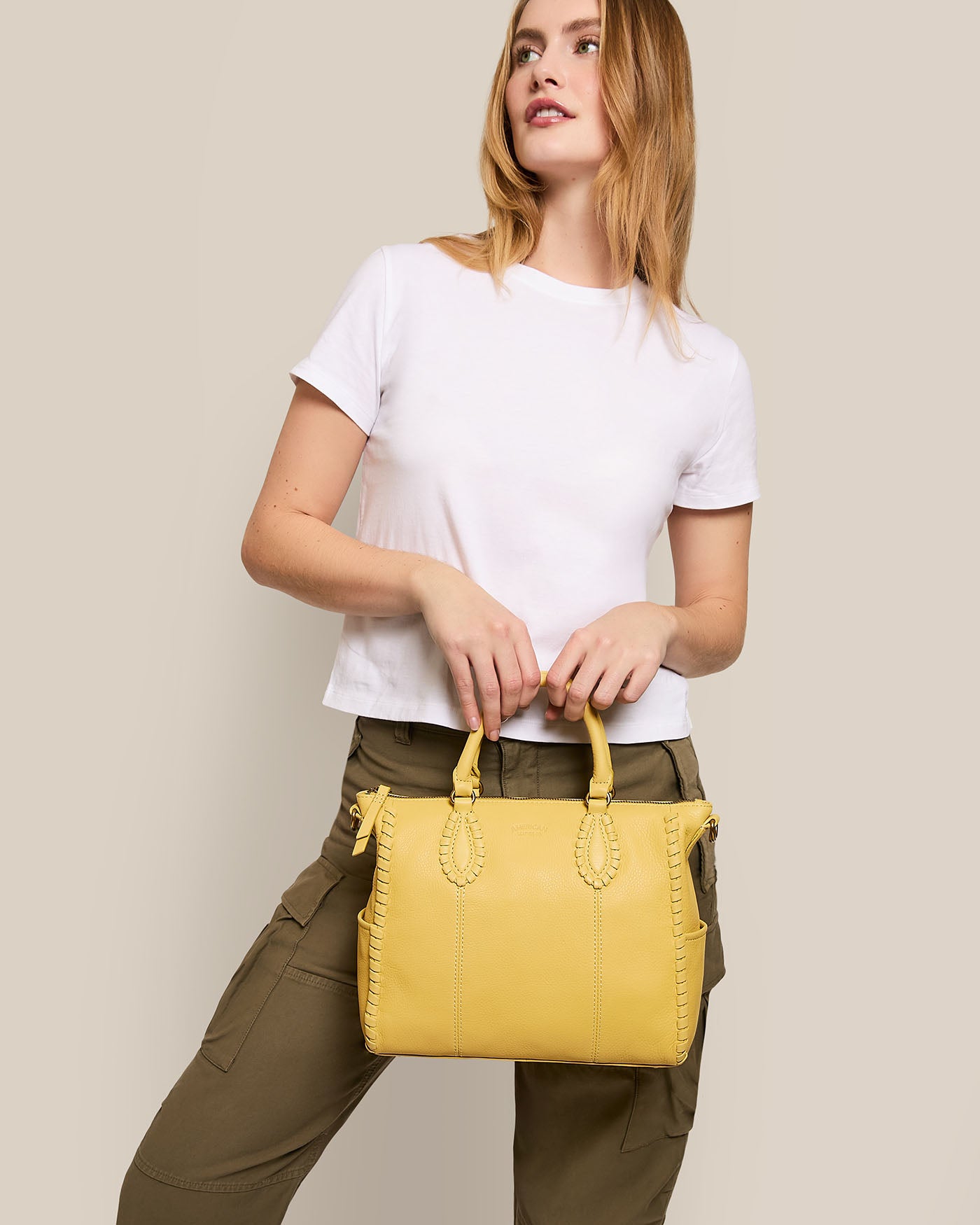 A person in a white t-shirt and olive green pants holds an American Leather Co. Madison Satchel with hand-woven glove leather detailing against a simple beige background.