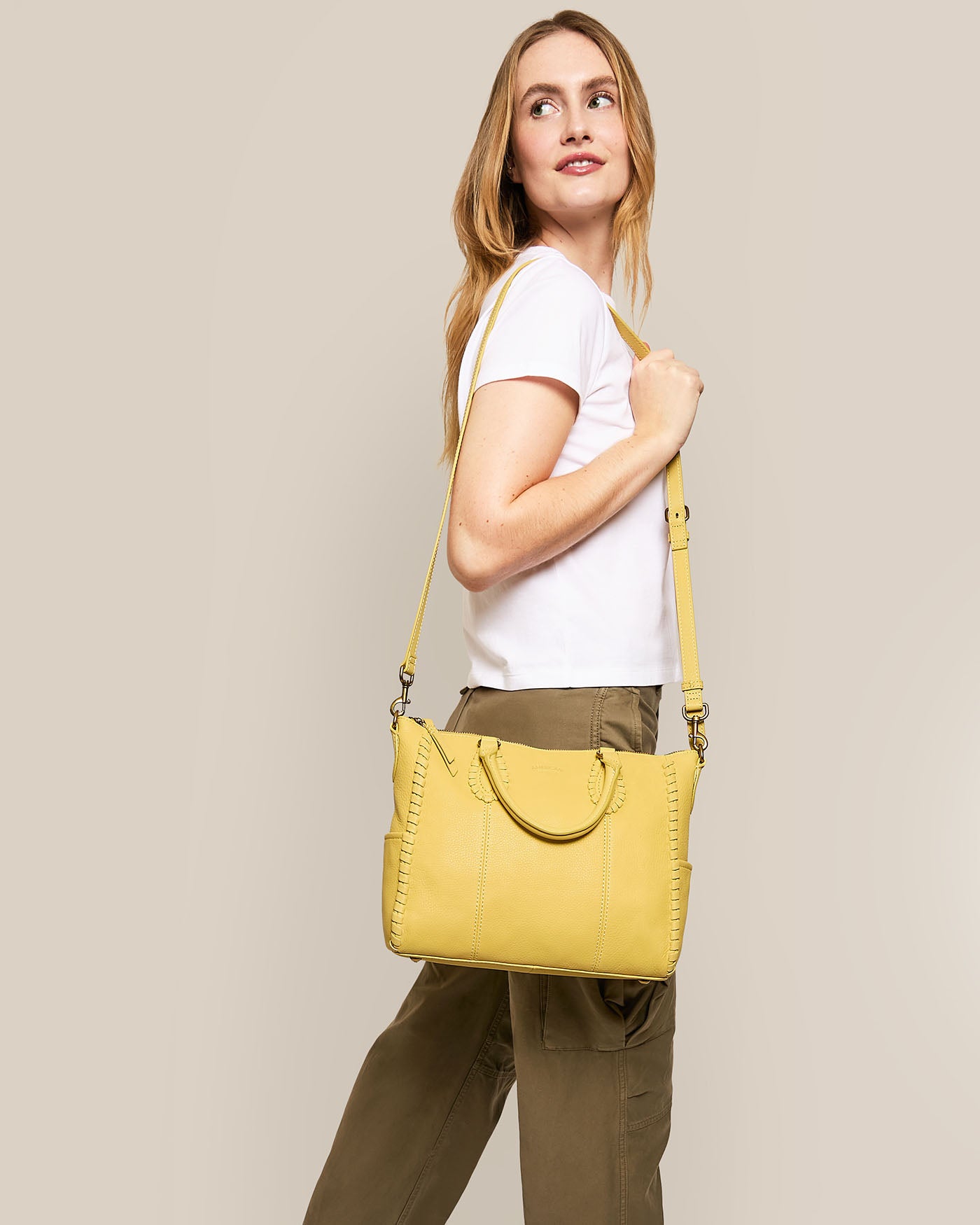 A person with long hair smiles while carrying an American Leather Co. Madison Satchel, showcasing exquisite hand-woven detailing. They wear a white t-shirt and olive green cargo pants against a plain beige background.