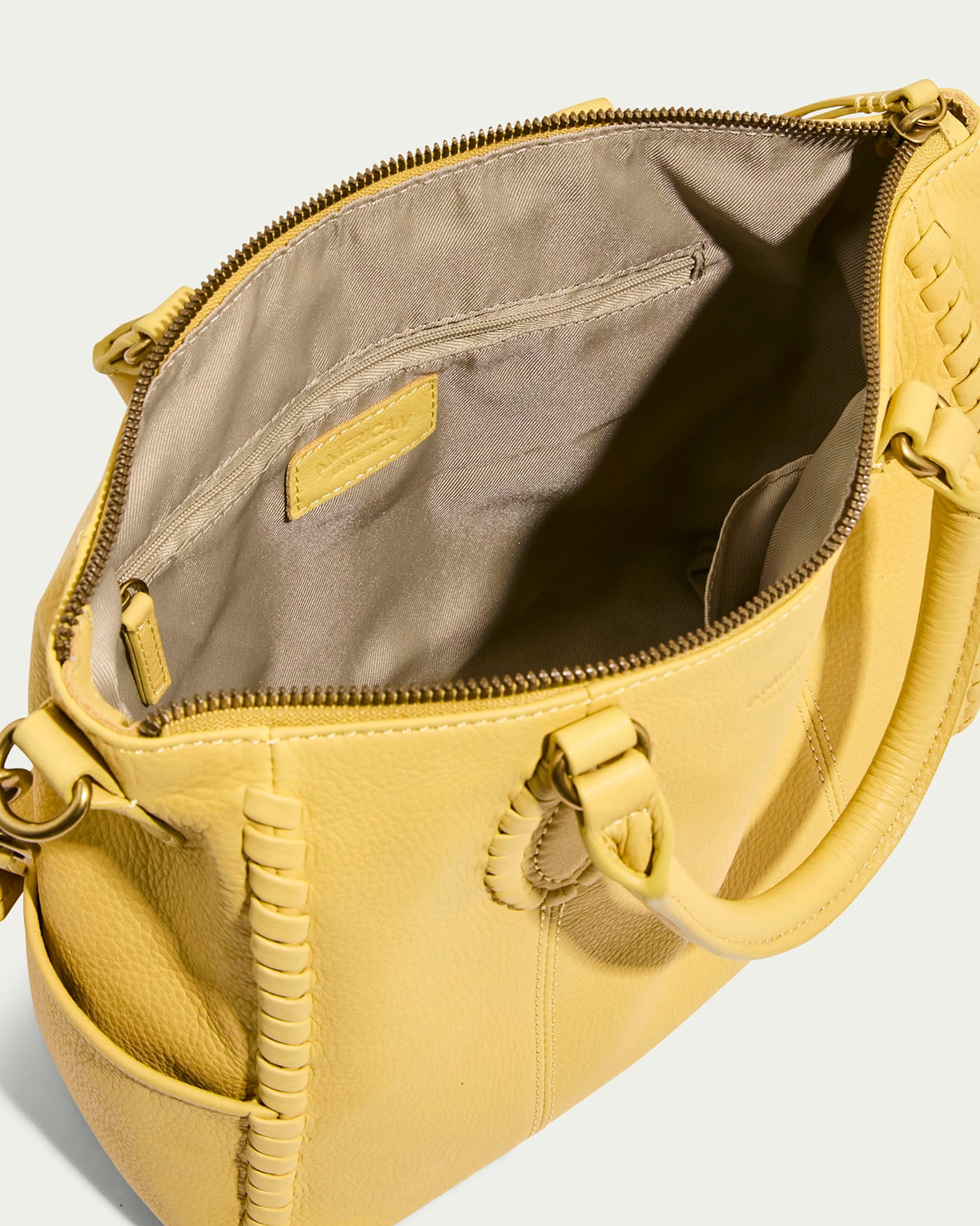 The Madison Satchel by American Leather Co. is a yellow leather handbag featuring visible stitching and a textured design. Made from glove leather, it boasts hand-woven detailing, an open top with a spacious interior, zip pocket, fabric lining, zip closure, and sturdy handles.