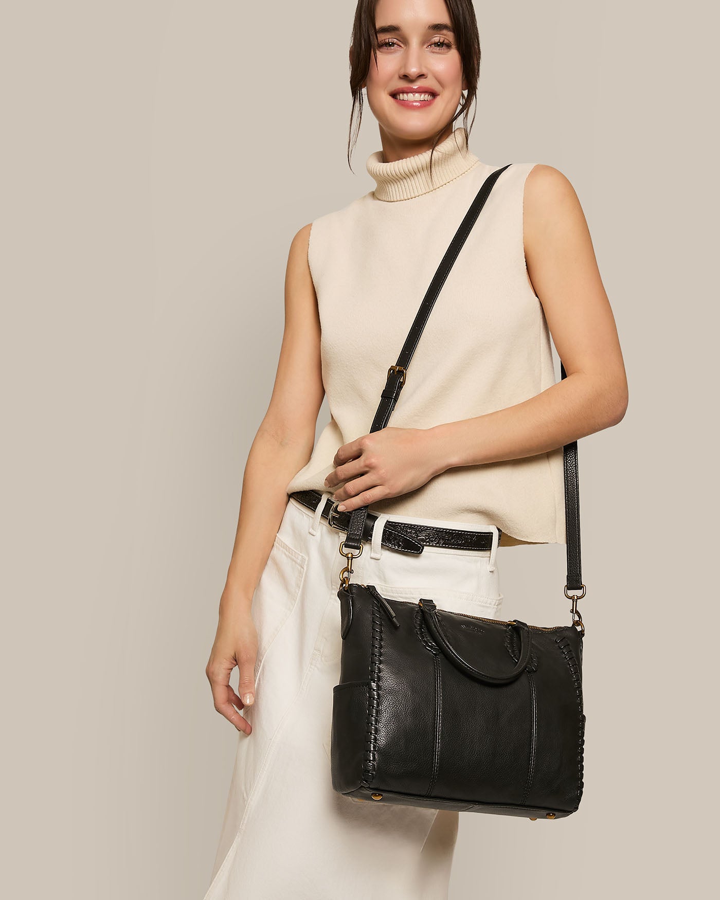 A smiling woman in a sleeveless turtleneck and white pants holds an American Leather Co. Madison Satchel, featuring black glove leather and hand-woven detailing, against a neutral background.