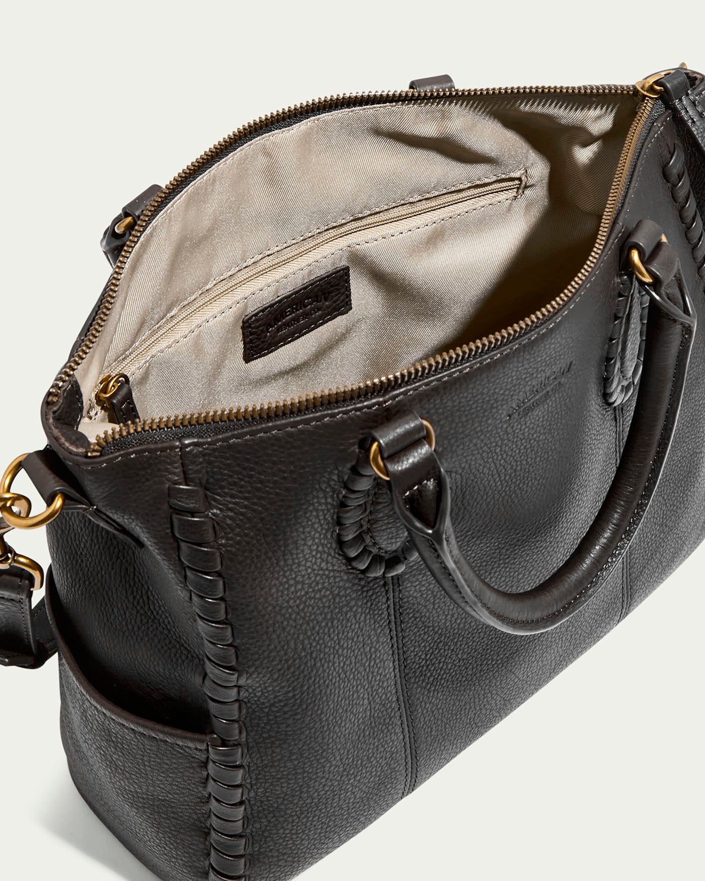 The Madison Satchel by American Leather Co. is crafted from glove leather with hand-woven details and gold hardware. Inside, a beige fabric lining and zippered pocket await. This black handbag has two handles and an adjustable shoulder strap for versatile use.