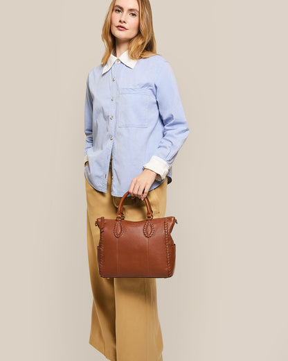 A woman stands against a plain beige background, wearing a light blue button-up shirt and tan wide-leg pants. She holds an American Leather Co. Madison Satchel, known for its glove leather and exquisite hand-woven detailing.