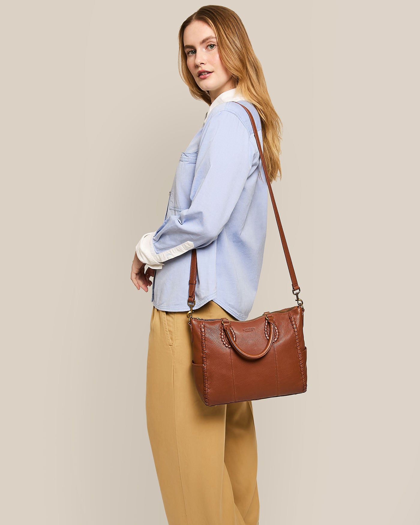 A person in a light blue button-up shirt and beige pants stands sideways against a plain background, holding the American Leather Co. Madison Satchel with glove leather and hand-woven detailing over their shoulder.