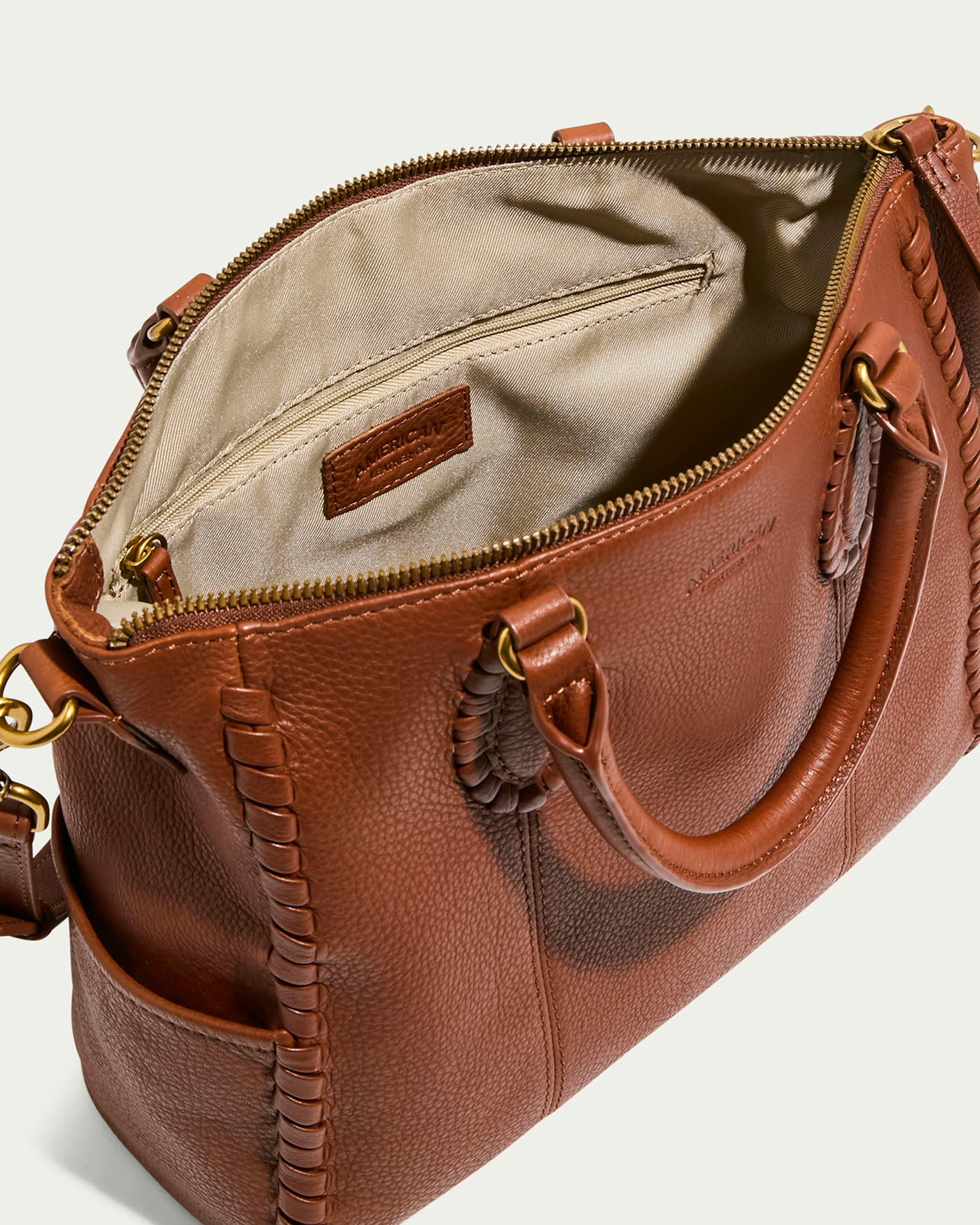 A brown Madison Satchel by American Leather Co. crafted from glove leather with hand-woven detailing and a zipper, shown from above. It features an open design with a beige interior, zippered pocket, brand label, handles, and an adjustable shoulder strap with brass clasps.