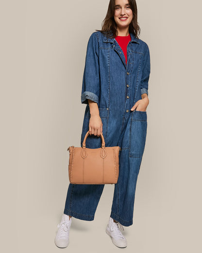 A woman in a denim jumpsuit and white sneakers holds an American Leather Co. Madison Satchel with hand-woven detailing, smiling against a plain background with her right hand in her pocket.