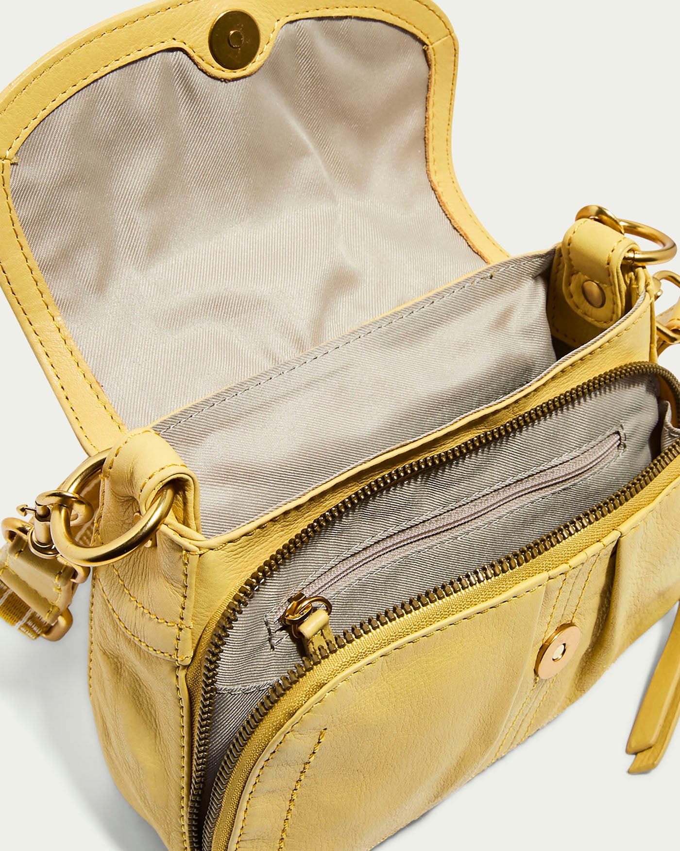 The American Leather Co. Marino Mini Crossbody is a small yellow glove leather shoulder bag with an open flap, magnetic closure, zippered compartment, and adjustable strap with metal hardware. It features a light beige fabric lining inside.