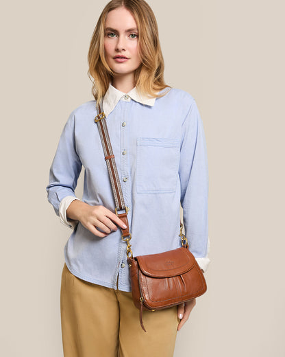 A woman with medium-length blonde hair stands against a beige background wearing a light blue button-up shirt and beige pants, holding the Marino Mini Crossbody by American Leather Co., crafted from glove leather and featuring a detachable strap.