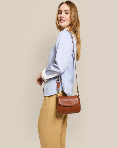 A person with long hair stands against a beige background, smiling and wearing a light blue shirt with beige pants. They're carrying the Marino Mini Crossbody by American Leather Co., made from glove leather with a detachable strap, while looking over their shoulder.