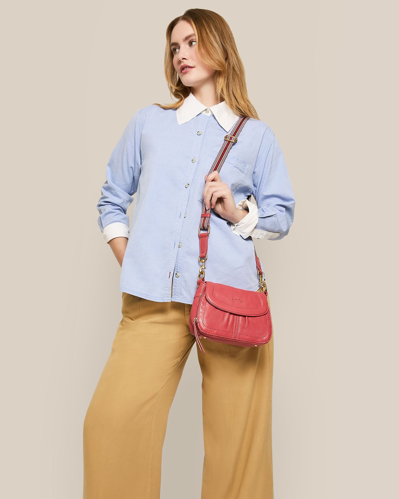 A person with long hair poses against a plain background, wearing a light blue button-up shirt and tan wide-leg pants. They carry the Marino Mini Crossbody by American Leather Co., a pink bag with an adjustable strap, over their shoulder, gazing confidently to the side.