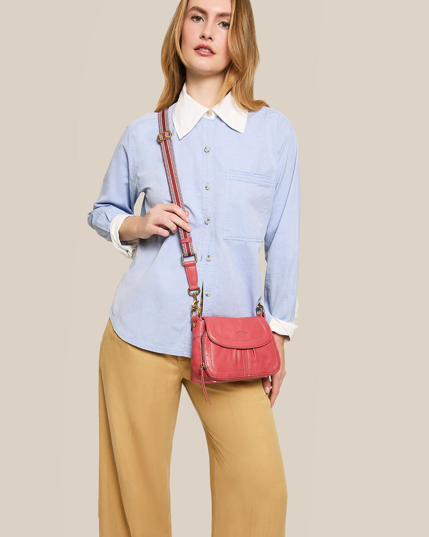 A person with long hair wearing a light blue button-up shirt with white collar and cuffs, beige pants, and holding the Marino Mini Crossbody bag from American Leather Co., which features an adjustable strap. The background is a plain neutral color.