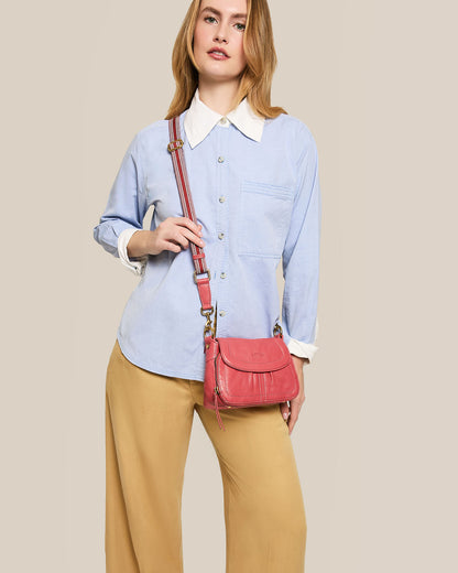 A person with long hair wearing a light blue button-up shirt with white collar and cuffs, beige pants, and holding the Marino Mini Crossbody bag from American Leather Co., which features an adjustable strap. The background is a plain neutral color.