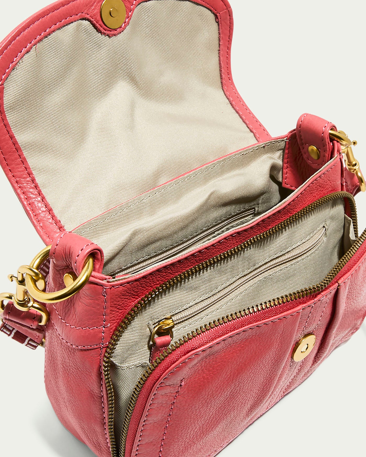 The Marino Mini Crossbody by American Leather Co. is crafted from pink glove leather, featuring a beige interior beneath its open flap. It includes a front zippered pocket with gold-toned hardware and button closure, plus an adjustable strap for versatility.