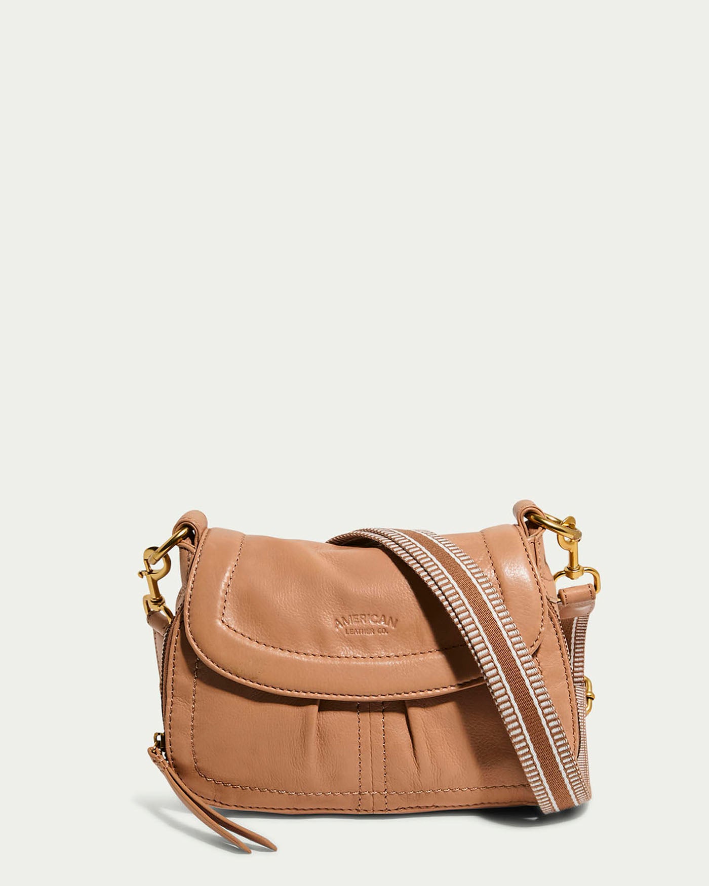 The Marino Mini Crossbody by American Leather Co. is a compact bag with a nutmeg leather exterior, flap closure, and pleated detailing. It has an adjustable striped strap with gold hardware and "American Leather Co." embossed on the front.