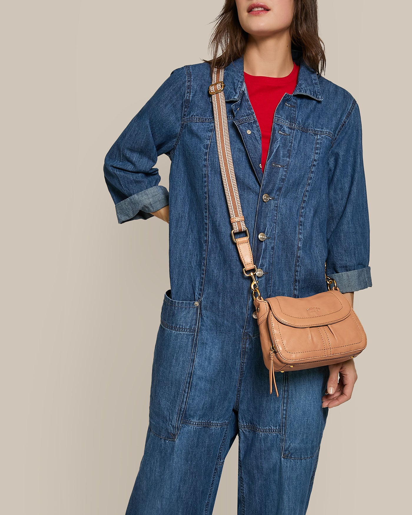 A person in a denim jumpsuit over a red top stands against a plain background, with American Leather Co.'s Marino Mini Crossbody featuring a brown strap on their shoulder, highlighting its compact design. Their left hand is in the jumpsuit's pocket.