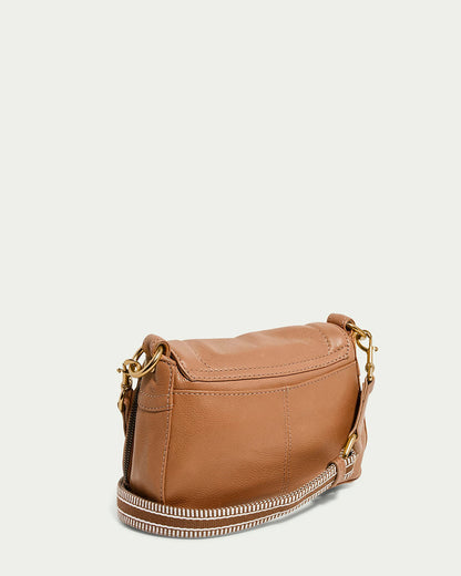 The Marino Mini Crossbody by American Leather Co. is a compact, light brown leather handbag with a flap closure and gold-tone buckle. It features a patterned, adjustable shoulder strap with gold clasps against a solid off-white background.