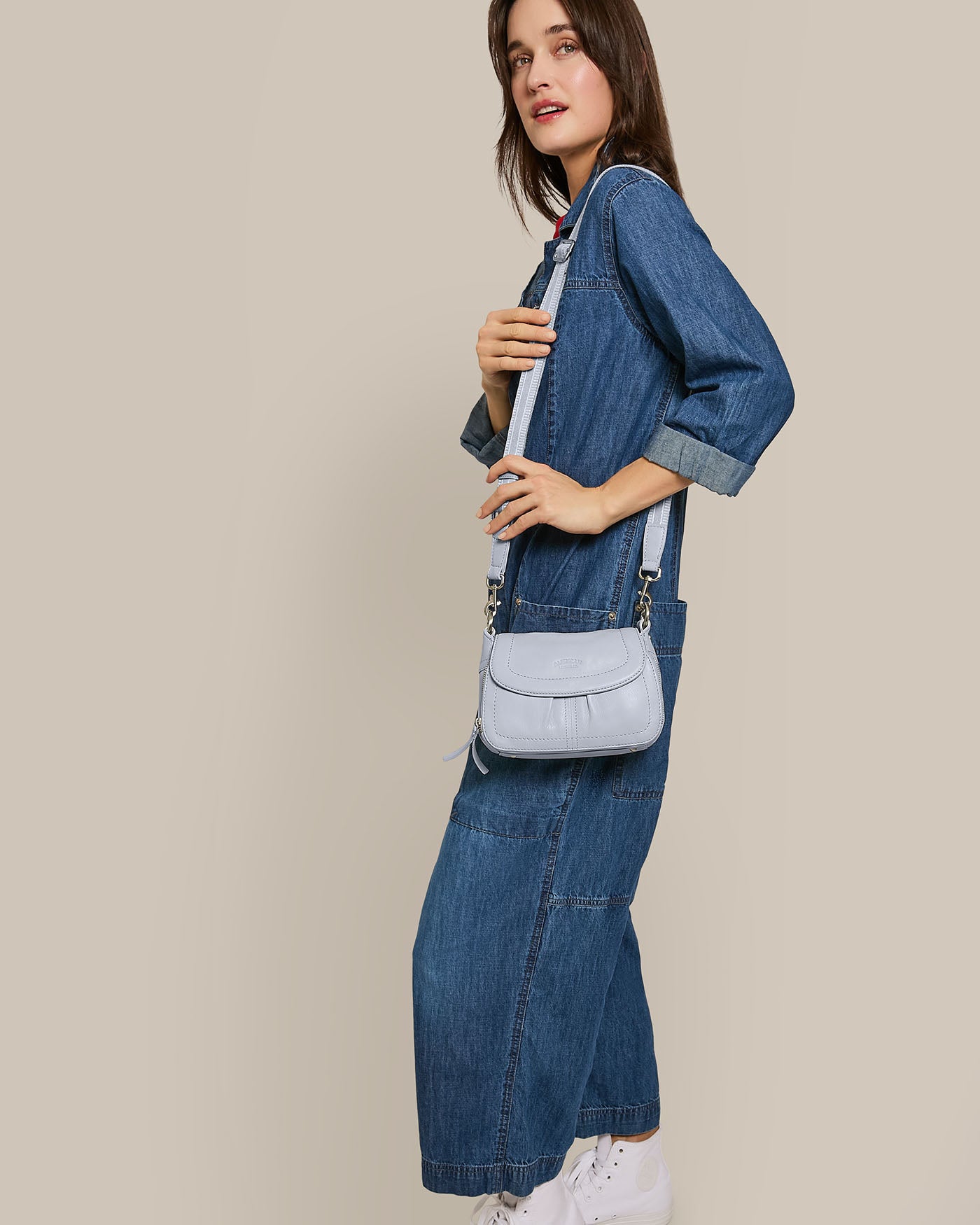 Wearing a denim jumpsuit and white sneakers, a person poses with the Marino Mini Crossbody featuring an adjustable strap from American Leather Co., set against a beige backdrop.