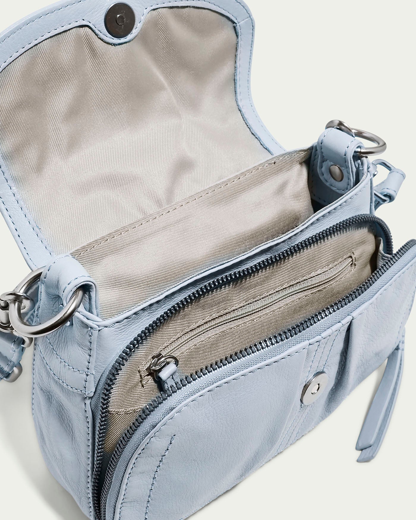 The Marino Mini Crossbody by American Leather Co. is a light blue bag with an open flap revealing a beige interior and zippered pocket. Made from glove leather, it includes side metal rings for an adjustable strap, allowing versatile wear.