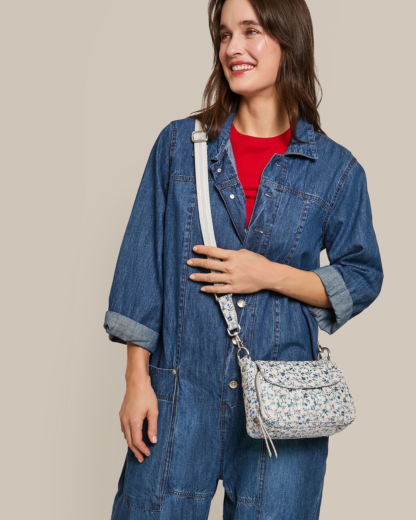 A person in a denim jumpsuit and red shirt smiles while holding the Marino Mini Crossbody from American Leather Co. Their long brown hair contrasts with the beige background, highlighting the handbag's compact and elegant design.